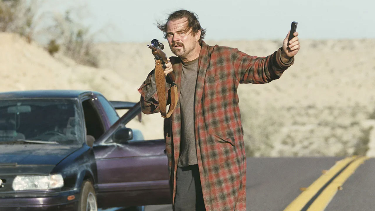 A rugged man (Leonardo DiCaprio) in a plaid coat aims a rifle while holding a handgun on an empty desert highway beside a stopped car in a tense scene from One Battle After Another.