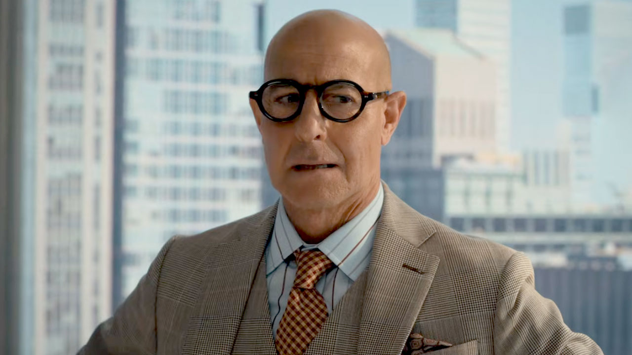 Stanley Tucci in a scene from The Devil Wears Prada 2, wearing glasses and a tan suit with a city skyline in the background.