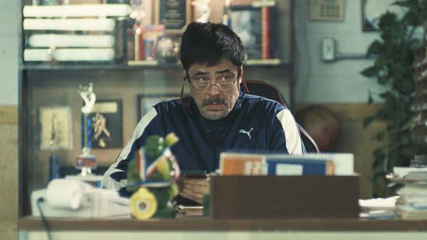 Actor Benicio del Toro wearing glasses and sitting at a disheveled desk in One Battle After Another