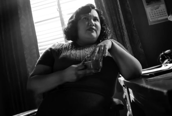 Karen Rodriguez as Janet in Spider-Noir, seated in a black-and-white office setting while holding a small container.