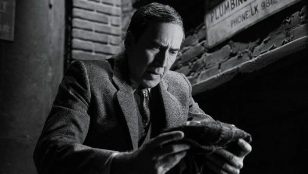 Nicolas Cage sits in a dark, black-and-white noir setting, examining a mask in his hands.