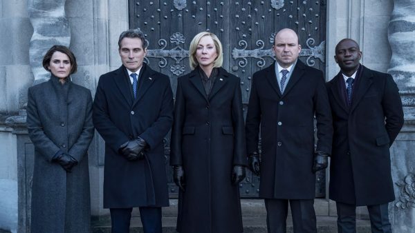 Keri Russell, Rufus Sewell, Allison Janney, Rory Kinnear and David Gyasi from Netflix’s ‘The Diplomat’ stand outside a large stone building in formal coats.